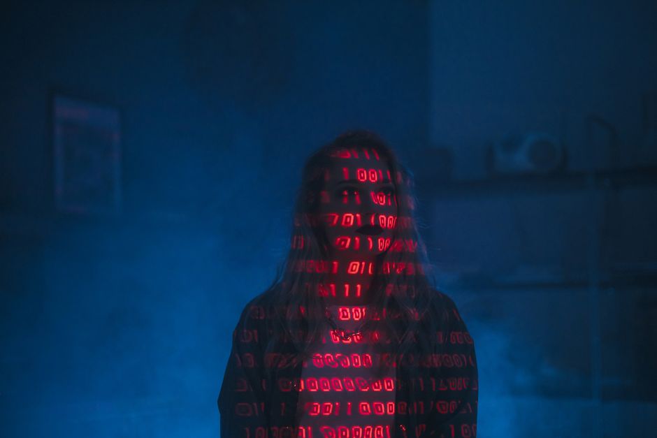 A person standing in a dimly lit room with a dark background, their face obscured by a hood. Red digital code or binary numbers are projected onto their face and upper body, creating a futuristic, cyb