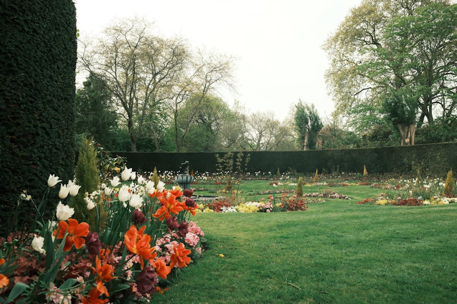 A well-maintained garden featuring a lush, neatly trimmed green lawn bordered by vibrant flower beds with a variety of blooming plants in shades of white, orange, pink, and purple. On the left, a tall