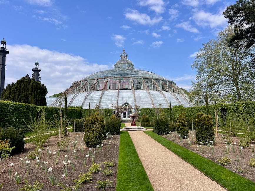 A well-maintained garden featuring a lush, neatly trimmed green lawn bordered by vibrant flower beds with a variety of blooming plants in shades of white, orange, pink, and purple. On the left, a tall