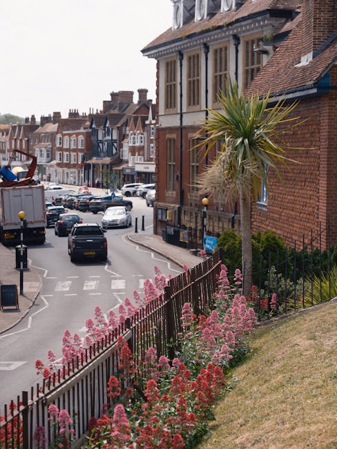 A residential street in Muswell Hill featuring a small garden area with pink and purple flowering plants along a wrought iron fence. Behind the garden, there is a tall, slender palm tree with long, sp